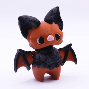 May include: A small, orange and black bat figurine with large eyes and a pink nose. The bat has wings that are spread out.