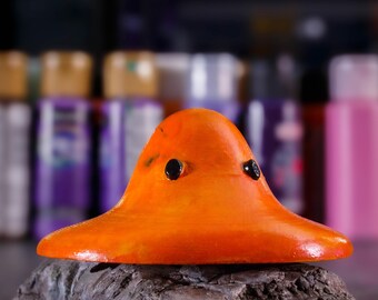 May include: Orange ceramic mushroom figurine with black eyes. The mushroom is sitting on a gray rock.