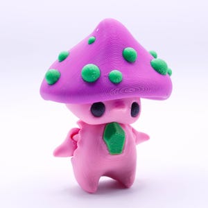 May include: A pink and purple mushroom-shaped figurine with green spots. The figurine has a small, round face with black eyes and a green gem on its chest.