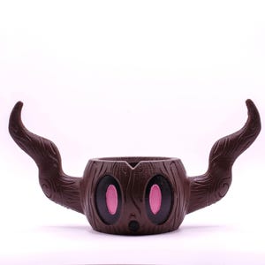 May include: A brown ceramic planter shaped like a tree stump with two long, curved branches. The planter has a cartoon face with two large pink eyes and a small brown mouth.