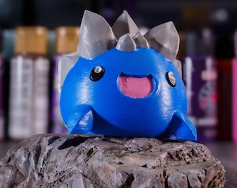 May include: A blue and grey 3D printed figurine of a smiling, cartoon-like creature with spikes. The creature is sitting on a grey rock.