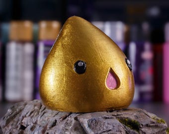 May include: A gold-colored, cartoon-like figurine with a pink nose and black eyes. The figurine is sitting on a rock-like surface.