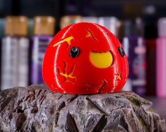 May include: A red and yellow 3D printed figurine of a cartoon character with a smiling face and black eyes. The figurine is sitting on a gray rock.