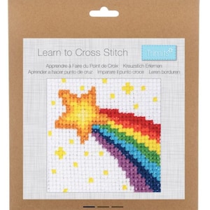 Op de afbeelding: Een Learn to Cross Stitch pakket van Trimits in bruine verpakking. De voorzijde toont een kruissteekpatroon van een gele ster met een regenboogkleurige staart op een wit raster, omringd door kleine gele sterretjes op een witte ondergrond.