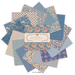 May include: A stack of square fabric swatches arranged in a starburst pattern. The fabrics feature various blue, cream, and pink patterns including floral prints, polka dots, stripes, and gingham. A central paper band reads Creating Memories.