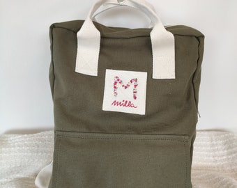 Könnte beinhalten: Ein grüner Canvas-Rucksack mit weißen Trägern und einem kleinen gestickten Aufnäher mit der Aufschrift "milla" und einem floralen "M" in der Mitte.