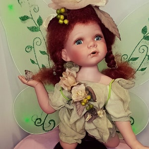 Może przedstawiać: Porcelanowa lalka z czerwonymi włosami, ubrana w zieloną sukienkę i skrzydła wr&oacute;żki. Lalka trzyma w ręku małego ptaka i ma na głowie wianek z kwiat&oacute;w.