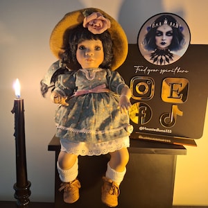 May include: A porcelain doll with a floral dress and a straw hat sits on a wooden surface. The doll has long black hair and brown eyes. A lit candle is in the background. The doll is wearing white socks and brown shoes. The doll is sitting in front of a black sign with gold social media icons and the text "Find your spirit here @Hauntedluna333".