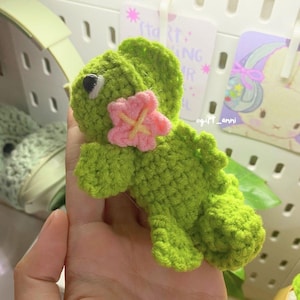 Könnte beinhalten: Ein handgefertigtes grünes Häkeldinosaurier mit einer rosa Blumenverzierung. Der Dinosaurier hat eine detaillierte, strukturierte Optik und ein kleines schwarzes Auge. Das Design ist skurril und charmant, perfekt für Kinderspielzeug oder Raumdekoration.