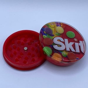 以下が含まれることがあります： スッキルズのキャンディーデザインが施された赤と透明なプラスチック製のグラインダー。グラインダーの上部は、スッキルズのデザインが施された透明な蓋になっており、グラインダーの下部は、グラインダーが付いた赤いベースになっています。