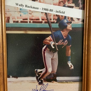 May include: A framed photograph of a baseball player in a blue and orange uniform swinging a bat. The player is wearing a white helmet with a blue logo. The photo is signed "Wally Backman" and includes the text "Wally Backman - 1980-88 - infield".