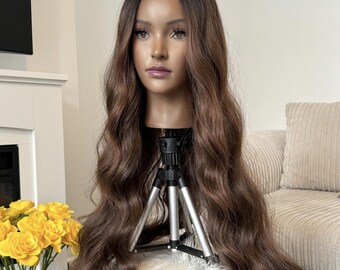 May include: A long, wavy, brown wig with lighter brown highlights. The wig is on a mannequin head and is styled in loose waves. The wig is sitting on a gray stool with a white fur throw on top.