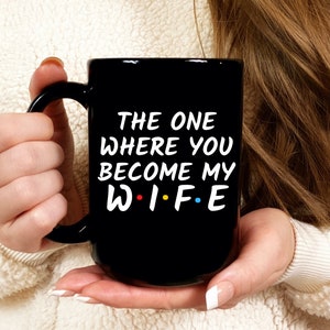 Op de afbeelding: Zwarte keramische mok met de witte tekst "THE ONE WHERE YOU BECOME MY W.I.F.E", waarbij het woord "WIFE" in de stijl van de tv-serie "Friends" is. De mok heeft een groot handvat.