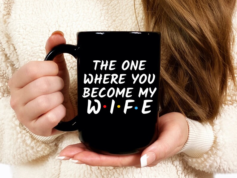 Op de afbeelding: Zwarte keramische mok met de witte tekst "THE ONE WHERE YOU BECOME MY W.I.F.E", waarbij het woord "WIFE" in de stijl van de tv-serie "Friends" is. De mok heeft een groot handvat.