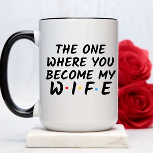Op de afbeelding: Witte keramische mok met een zwarte handgreep en rand. De mok heeft de tekst "THE ONE WHERE YOU BECOME MY WIFE" in het zwart, met het woord "WIFE" in de kleuren rood, geel, blauw en groen. De mok staat op een wit oppervlak.