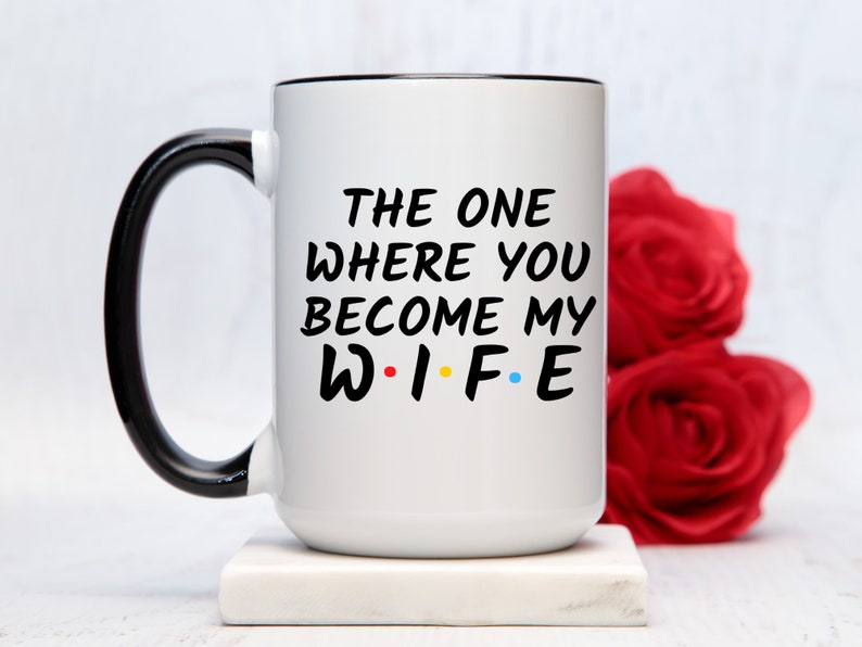 Op de afbeelding: Witte keramische mok met een zwarte handgreep en rand. De mok heeft de tekst "THE ONE WHERE YOU BECOME MY WIFE" in het zwart, met het woord "WIFE" in de kleuren rood, geel, blauw en groen. De mok staat op een wit oppervlak.