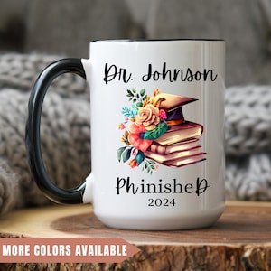 May include: White ceramic coffee mug with a black handle. The mug has a black rim and a design that says "Dr. Johnson Phinished 2024" with a graduation cap and flowers.