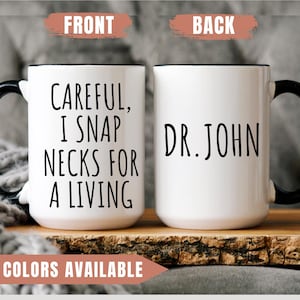 May include: White ceramic coffee mug with a black handle and black rim. The front of the mug reads "Careful, I snap necks for a living". The back of the mug reads "Dr. John".