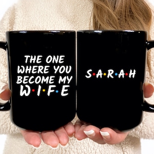 Op de afbeelding: Twee zwarte keramische mokken met witte tekst. Op de ene mok staat "THE ONE WHERE YOU BECOME MY WIFE" en op de andere "SARAH" met gekleurde stippen tussen de letters. De mokken hebben grote handvatten.