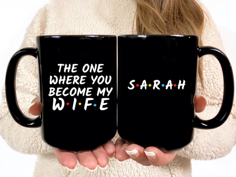 Op de afbeelding: Twee zwarte keramische mokken met witte tekst. Op de ene mok staat "THE ONE WHERE YOU BECOME MY WIFE" en op de andere "SARAH" met gekleurde stippen tussen de letters. De mokken hebben grote handvatten.