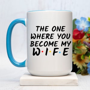 Op de afbeelding: Witte keramische mok met een blauw handvat en rand. De mok heeft de tekst "THE ONE WHERE YOU BECOME MY WIFE" in het zwart, met het woord "WIFE" in de stijl van de tv-serie Friends. Een leuk cadeau voor een bruiloft of verloving.
