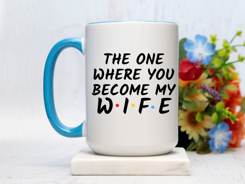 Op de afbeelding: Witte keramische mok met een blauw handvat en rand. De mok heeft de tekst "THE ONE WHERE YOU BECOME MY WIFE" in het zwart, met het woord "WIFE" in de stijl van de tv-serie Friends. Een leuk cadeau voor een bruiloft of verloving.
