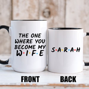 Op de afbeelding: Twee witte keramische mokken met zwarte handvatten en randen. Op &eacute;&eacute;n mok staat "THE ONE WHERE YOU BECOME MY WIFE" in zwarte tekst met kleurrijke stippen. De andere mok heeft de naam "SARAH" in kleurrijke tekst.