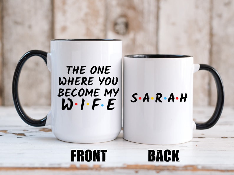 Op de afbeelding: Twee witte keramische mokken met zwarte handvatten en randen. Op &eacute;&eacute;n mok staat "THE ONE WHERE YOU BECOME MY WIFE" in zwarte tekst met kleurrijke stippen. De andere mok heeft de naam "SARAH" in kleurrijke tekst.