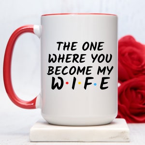 Op de afbeelding: Witte keramische mok met een rode handgreep en binnenrand. De mok heeft de zwarte tekst "THE ONE WHERE YOU BECOME MY WIFE", met het woord "WIFE" in de stijl van de tv-serie "Friends". Twee rode rozen op de achtergrond.