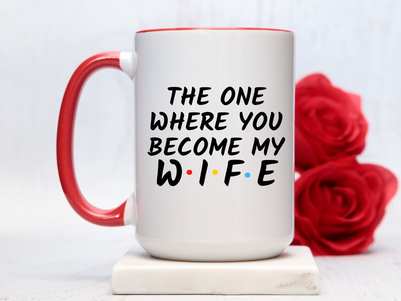 Op de afbeelding: Witte keramische mok met een rode handgreep en binnenrand. De mok heeft de zwarte tekst "THE ONE WHERE YOU BECOME MY WIFE", met het woord "WIFE" in de stijl van de tv-serie "Friends". Twee rode rozen op de achtergrond.