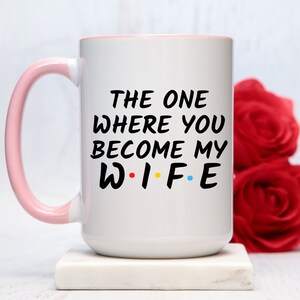 Op de afbeelding: Witte keramische mok met een roze handvat en rand. De mok heeft de tekst "THE ONE WHERE YOU BECOME MY WIFE" in het zwart, met de letters "WIFE" in de stijl van de tv-serie "Friends". Rode rozen staan op de achtergrond.