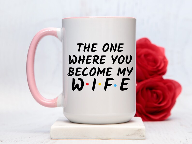 Op de afbeelding: Witte keramische mok met een roze handvat en rand. De mok heeft de tekst "THE ONE WHERE YOU BECOME MY WIFE" in het zwart, met de letters "WIFE" in de stijl van de tv-serie "Friends". Rode rozen staan op de achtergrond.