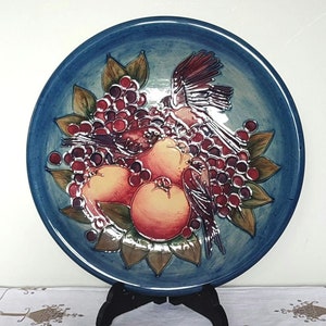 May include: A blue ceramic plate with a raised design of birds and fruit. The plate features a blue background with a raised design of two birds perched on a branch with red berries and pears. The birds are painted in shades of brown, red, and white.