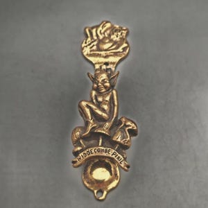 May include: A gold-coloured metal spoon with a pixie figure seated on a mushroom, and the words "Widecombe Pixie" inscribed on a banner. The spoon's handle has a decorative top with a cherub design.