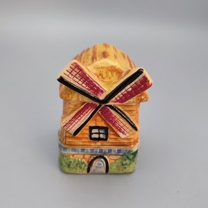May include: A ceramic windmill ornament with a thatched roof and red and black blades. The windmill has a rectangular base with a small arched doorway and a window. The colors are primarily yellow, brown, red, and green.