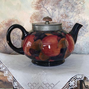 May include: A red and black teapot with a pewter lid and a knob on top. The teapot is decorated with a floral pattern and has a round body.