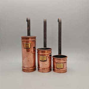 May include: A set of three cylindrical copper measuring cups with long, dark metal handles. Each cup features a small brass plaque on the front, with the tallest cup clearly marked with the text 1 PINT. The cups vary in height and width.