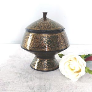 May include: A decorative, lidded container with a pedestal base. The container is painted in a dark green and gold floral pattern. The lid has a small, dark brown knob. A white rose is in the foreground.