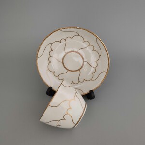 May include: A white ceramic teacup and matching saucer set featuring a delicate gold-coloured outline of a floral pattern. The saucer has a scalloped gold rim, and the teacup is positioned in front of the saucer, both displaying a consistent design.