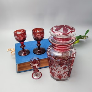 May include: Set of three ruby red glass decanters with etched floral designs. The decanters have a clear glass base and a red glass top. The decanters are on a blue book.