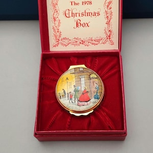 May include: A round, gold-toned compact with a painted scene of people walking in a snowy street. The compact is in a red velvet-lined box with a white label that reads "The 1978 Christmas Box".