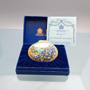May include: An oval Bilston & Battersea Enamels trinket box with a gold-tone rim, featuring a painted landscape with blue morning glory flowers and the year 2000. The box is presented in a dark blue velvet-lined box with a certificate of origin.