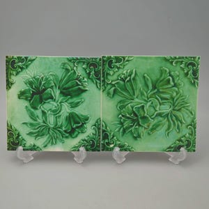 May include: Two square, green ceramic tiles with a floral relief design. The tiles feature a detailed depiction of flowers and foliage, with a darker green hue highlighting the raised elements. The corners of the tiles have decorative scrollwork.