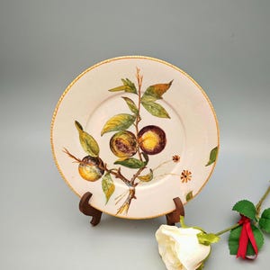 May include: A cream-coloured ceramic plate featuring a hand-painted design of plums on a branch with green leaves. The plate has a gold rim and is displayed on a wooden stand. A white rose with a red ribbon is in the foreground.