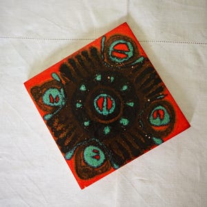 May include: Square ceramic tile with a vibrant design. The tile has a red border and a central pattern in shades of brown, teal, and red. The design features a circular motif with abstract shapes. The tile is likely for decorative use.