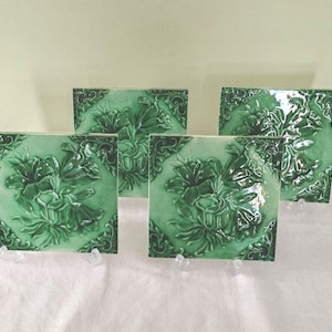 May include: Four square green ceramic tiles with a raised floral design. The tiles have a glossy finish and are displayed on clear stands. The floral pattern is detailed with leaves and petals, and the edges have a decorative border.
