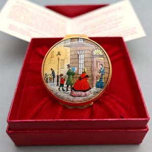 May include: A round gold-toned metal pill box featuring a colourful illustration of 19th-century figures on a street scene. The lid depicts people in period clothing near a building entrance. The box is presented inside a red velvet-lined box.