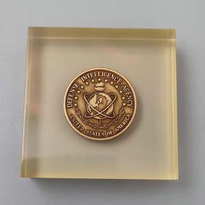 May include: A wooden plaque with a gold-coloured seal of the Defence Intelligence Agency. The seal features a globe with a torch above it, surrounded by stars and the words "Defence Intelligence Agency" and "United States of America".