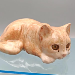 May include: A ceramic cat figurine in a resting, lying-down position. The sculpture features a glossy, mottled finish with shades of light orange, cream, and tan. The cat has large, dark, expressive eyes and pointed ears, with its front paws tucked.