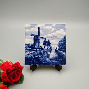May include: Square ceramic tile with a blue and white illustration of a Dutch windmill scene. The artwork shows a windmill, water, trees, and a bridge under a cloudy sky. The tile is displayed on a black stand.